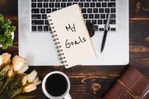My Goals written on a notepad sat ontop of a laptop keyboard. With autumnal flowers and a black coffee